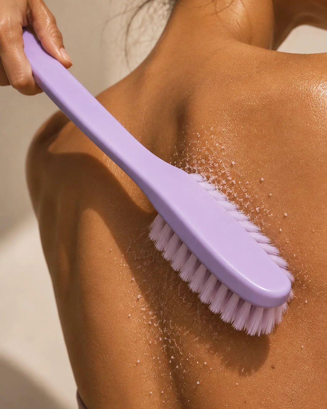 Close-up of a hand using a lavender-colored bristle brush to exfoliate the back and shoulder, with granular scrub particles visible scattering across sun-lit skin.